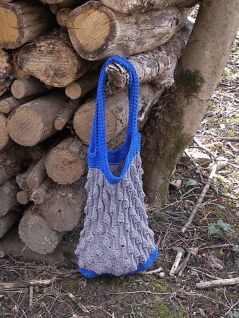 Gestrickte Beuteltasche mit breitem Träger, grauer Körper mit Reliefstruktur und blauen Einfassungen, hängt an einem Baumstamm vor Holzstapel
