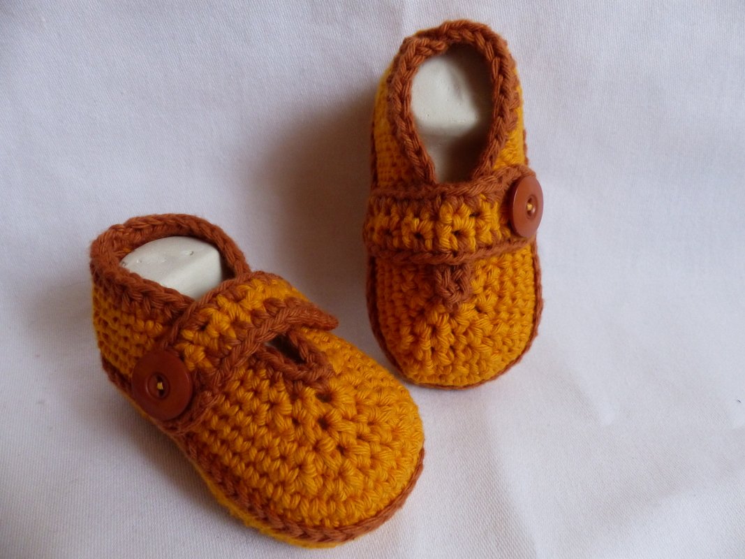 Zwei gehäkelte Babyschuhe in Orange und Braun mit Knopf auf weißem Hintergrund