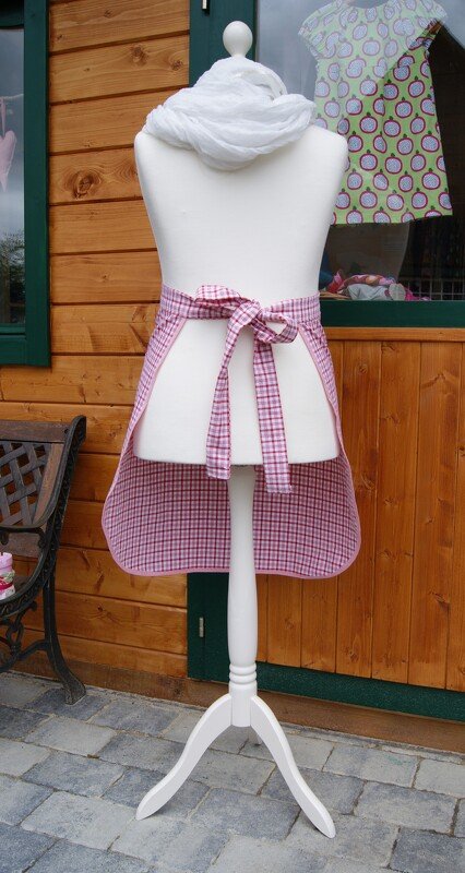 Weiße Schneiderpuppe mit rosa-rot karierter Halbschürze, hinten mit Schleife, vor einer Holzwand