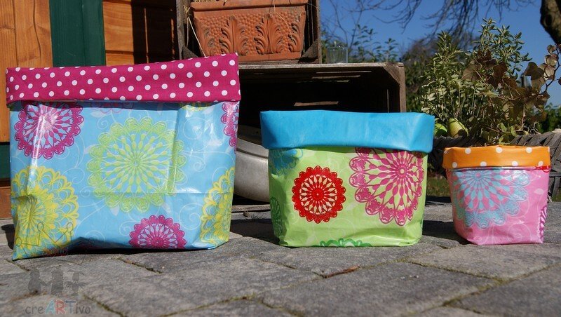 Drei bunte Utensilos aus Wachstuch in Blau, Grün und Rosa mit gemusterten Umschlägen auf Steinboden.