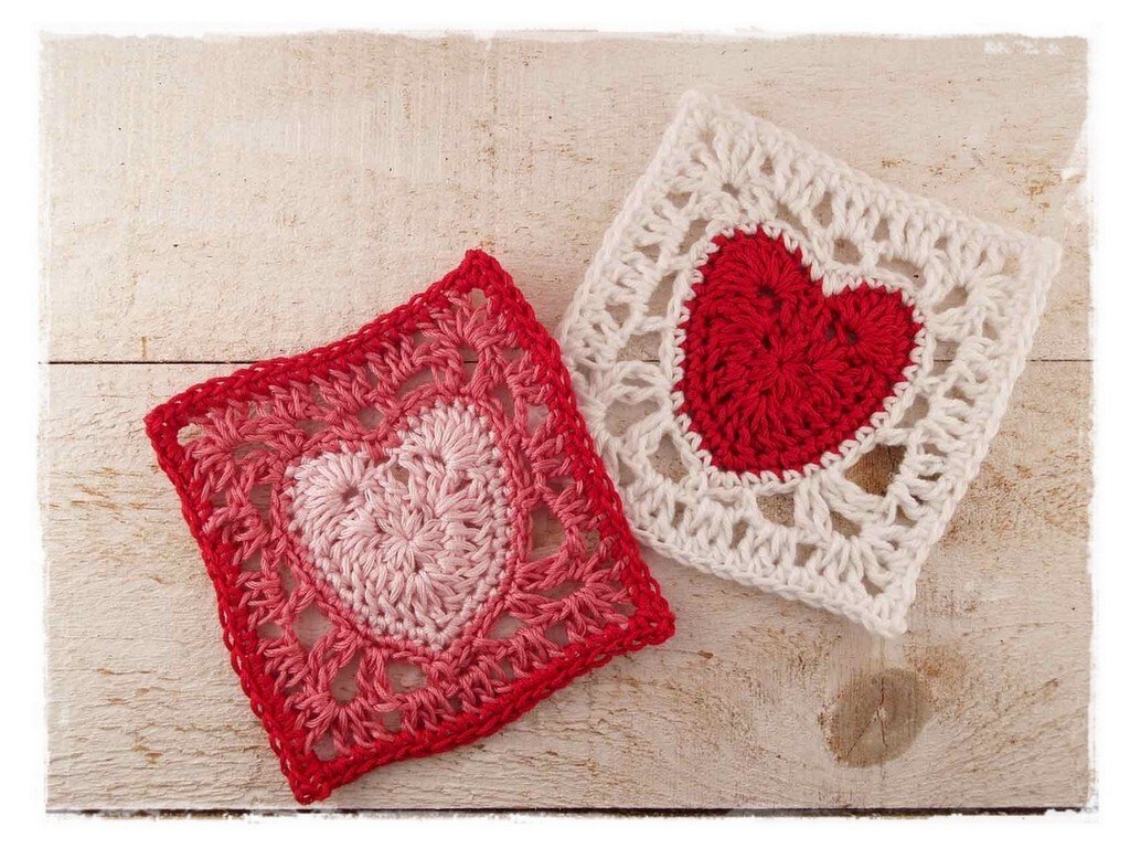 Zwei gehäkelte Granny Squares mit Herzmotiven in Rot, Rosa und Weiß auf hellem Holzuntergrund.
