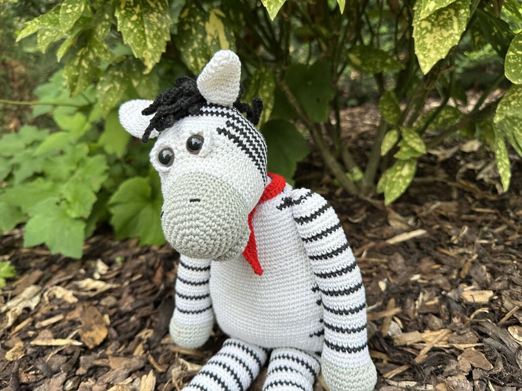 Gehäkeltes Zebra-Amigurumi mit rotem Halstuch auf Rindenmulch vor Pflanzen