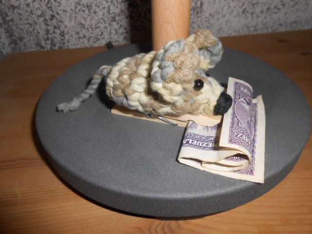 Gehäkelte Amigurumi‑Maus aus mehrfarbigem Garn auf einer Mausefalle neben gefaltetem Geldschein