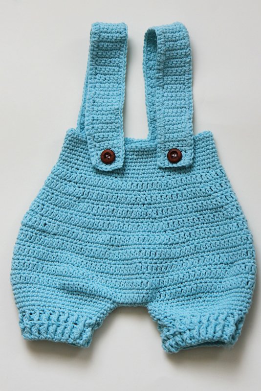 Hellblauer gehäkelter Baby-Strampler mit Trägern und dunklen Knöpfen, liegend.