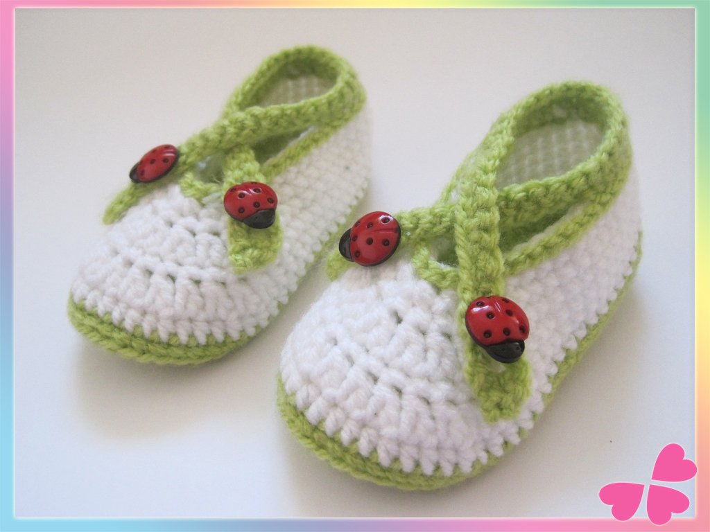Gehäkelte weiße Babyschuhe mit hellgrünen Riemchen und roten Marienkäfer-Knöpfen auf weißem Hintergrund.