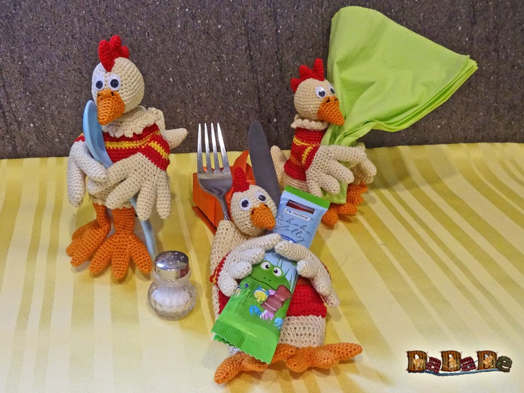 Drei gehäkelte Hühner halten Besteck, Serviette und Süßigkeit auf gelbem Tischläufer