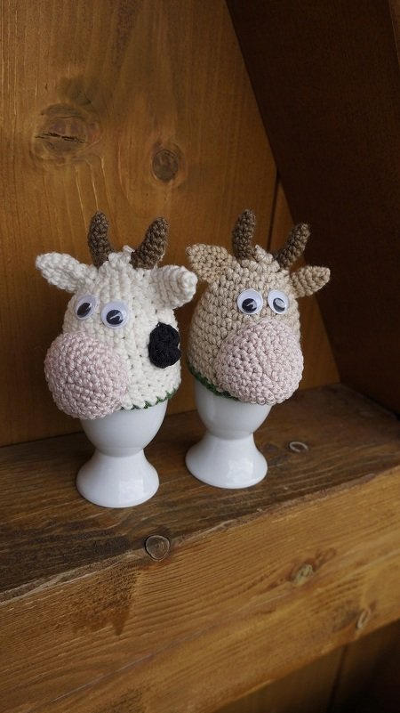 Zwei gehäkelte Kuh-Eierwärmer auf weißen Eierbechern, einer weiß mit schwarzem Fleck, der andere beige