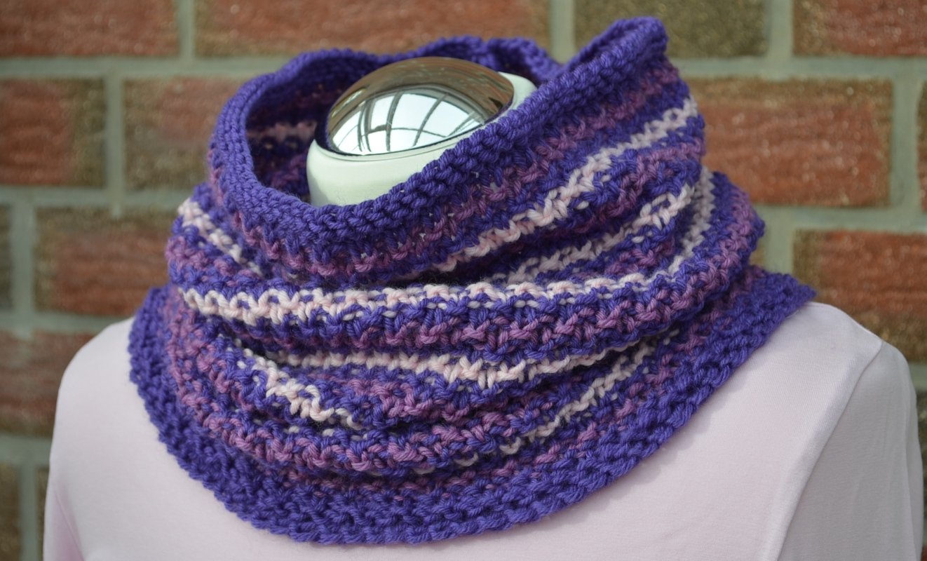 Gestrickter Loop‑Schal in Lila‑ und Rosatönen mit Streifen auf einer Büste.