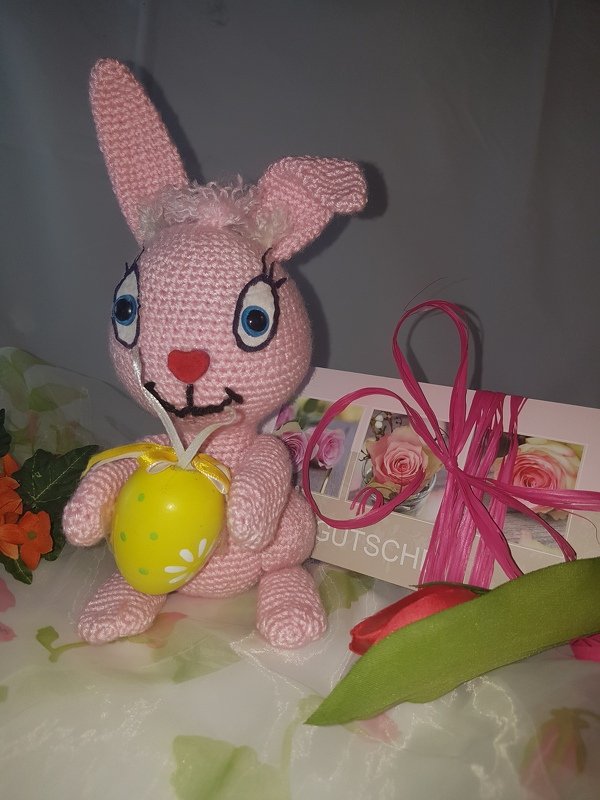 Rosa gehäkelter Amigurumi-Hase mit großen Augen, der ein gelbes Osterei mit weißen Punkten hält
