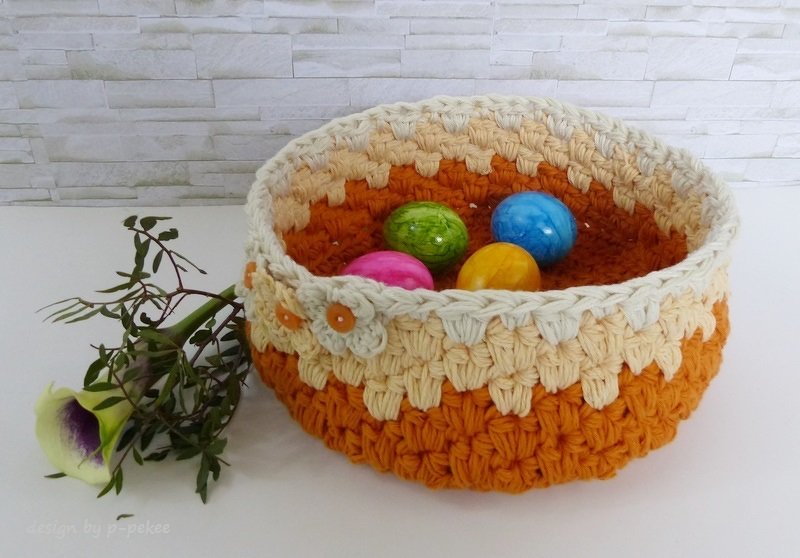 Gehäkelter runder Korb in Orange und Creme mit Blütenapplikation, gefüllt mit bunten Ostereiern.