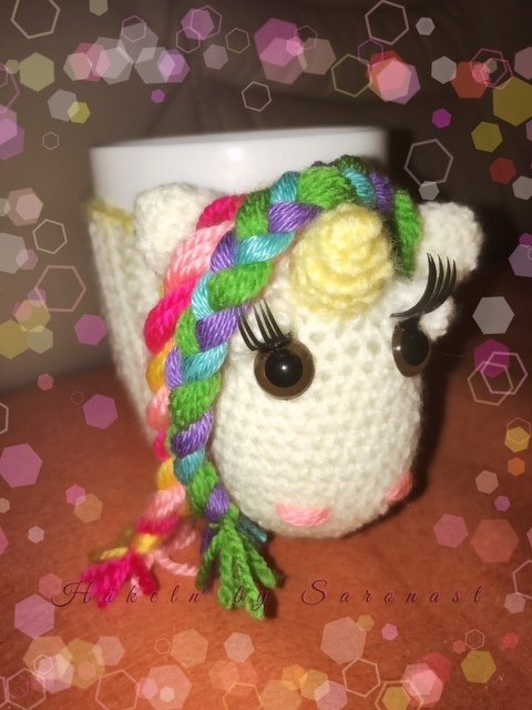 Gehäkelte cremeweiße Einhorn-Tassenhülle mit geflochtener Regenbogenmähne, gelbem Horn und Wimpern, umschließt eine weiße Tasse.