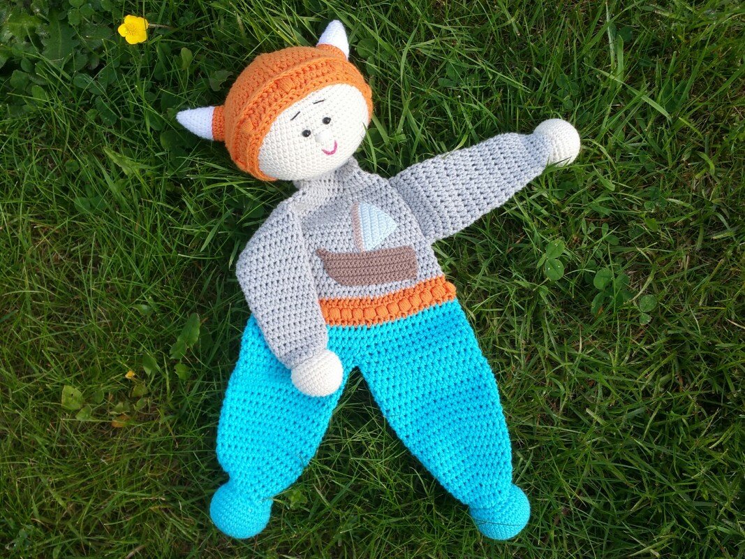 Gehäkeltes Schmusetuch im Wikinger-Look mit orangefarbenem Helm und hellblauer Hose auf einer grünen Wiese