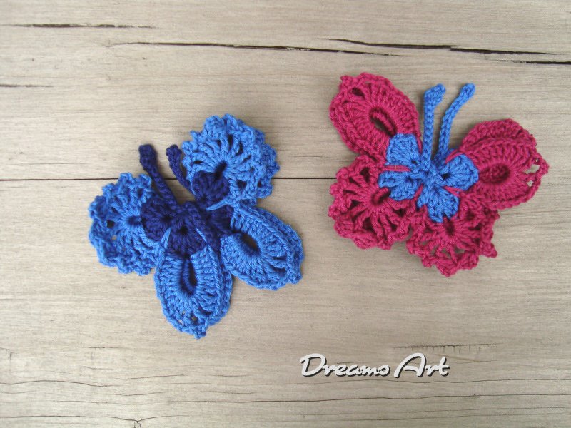 Zwei gehäkelte Schmetterlingsapplikationen, eine blau und eine pink, auf Holzuntergrund