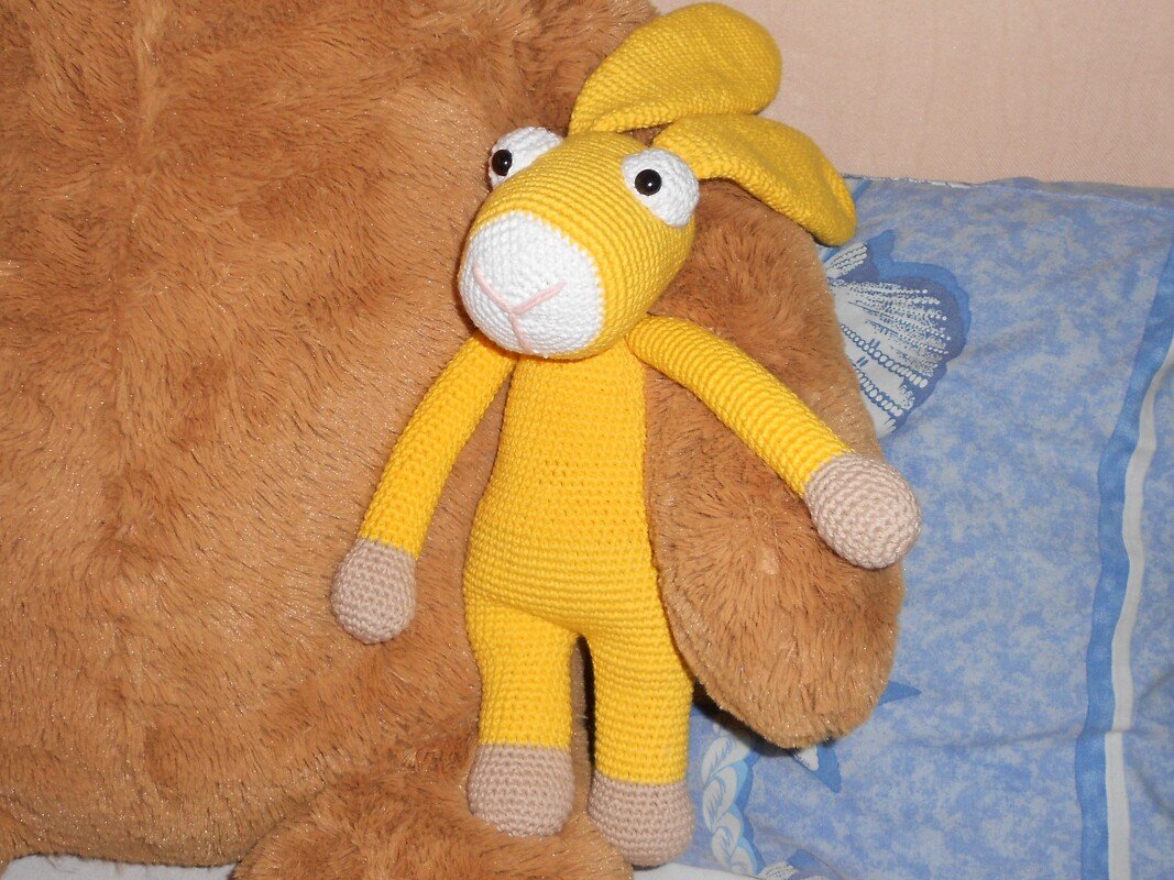 Gehäkelter gelber Amigurumi-Hase mit weißer Schnauze und beigen Pfoten, an braunem Kuscheltier lehnend