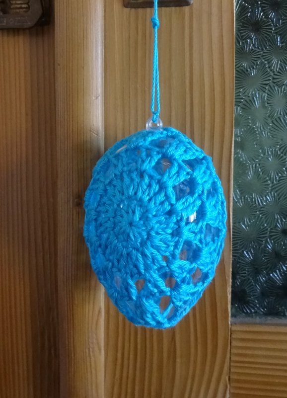 Türkises gehäkeltes Ei an einem Faden, hängend vor Holz und strukturierter Glasscheibe.