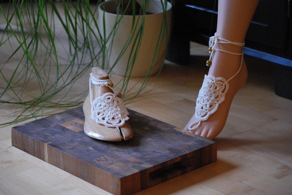 Cremefarbene gehäkelte Barfußsandalen, eine am Fuß, eine auf einer Schuhleiste, mit Holzperlen verziert.