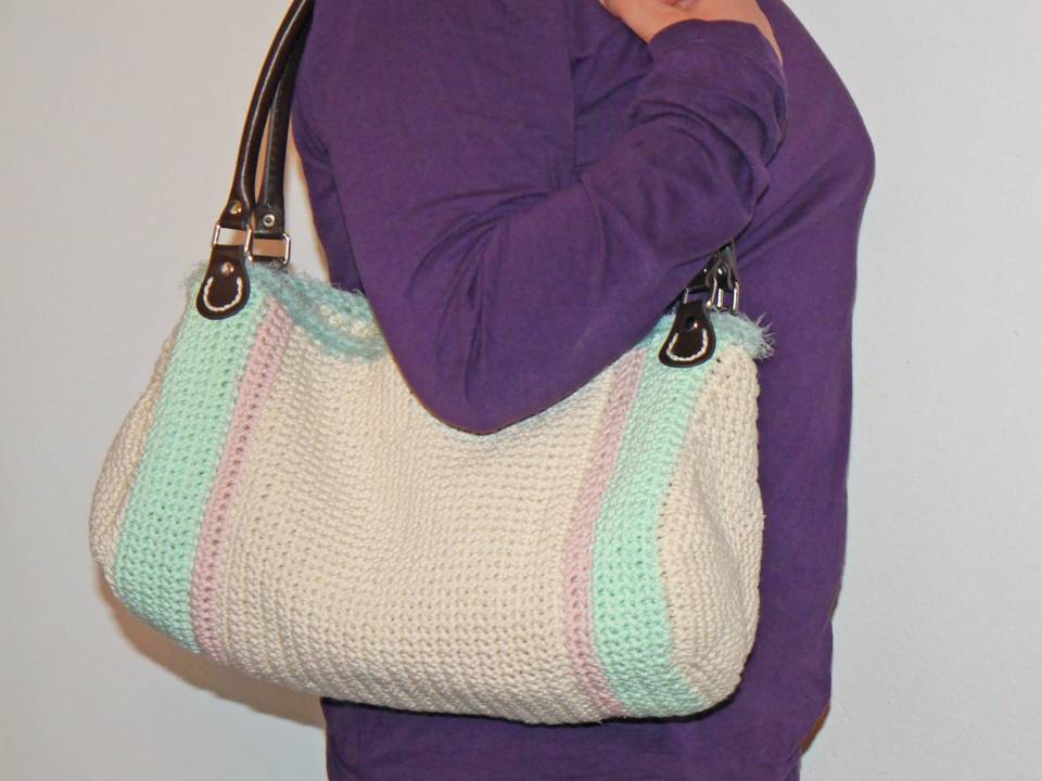 Gehäkelte Schultertasche creme mit mint- und rosafarbenen Streifen, braune Henkel, getragen an der Schulter einer Person in lila Oberteil