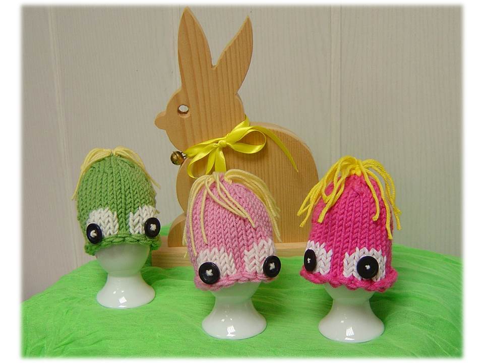 Drei gestrickte Eierwärmer in Grün, Rosa und Pink mit Knopfaugen und gelben Wollhaaren auf weißen Eierbechern, dahinter ein Holzhase.