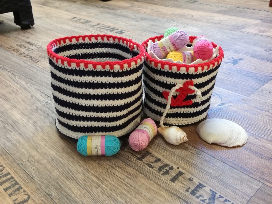 Zwei gehäkelte blau-weiß gestreifte Körbe mit roter Kante und Anker, daneben Garn und Muscheln