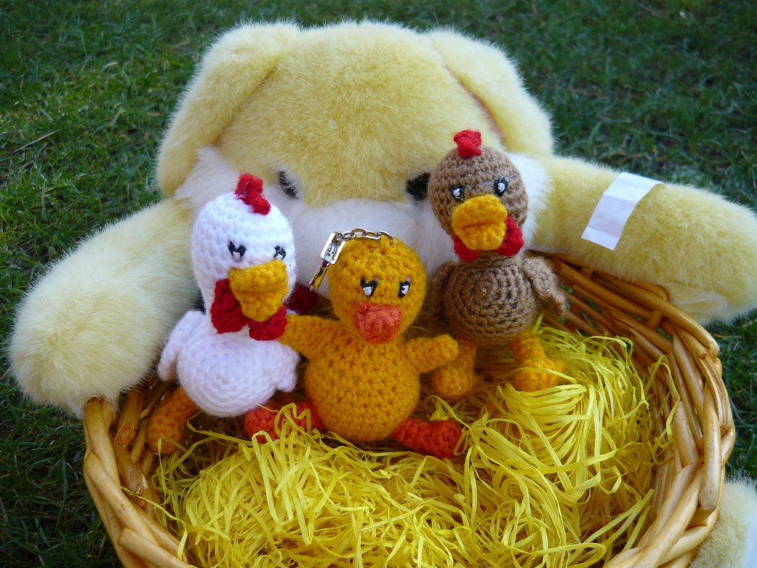 Drei gehäkelte Amigurumi-Vögel (zwei Hühner, ein Küken mit Schlüsselring) in einem Weidenkorb auf gelbem Stroh.
