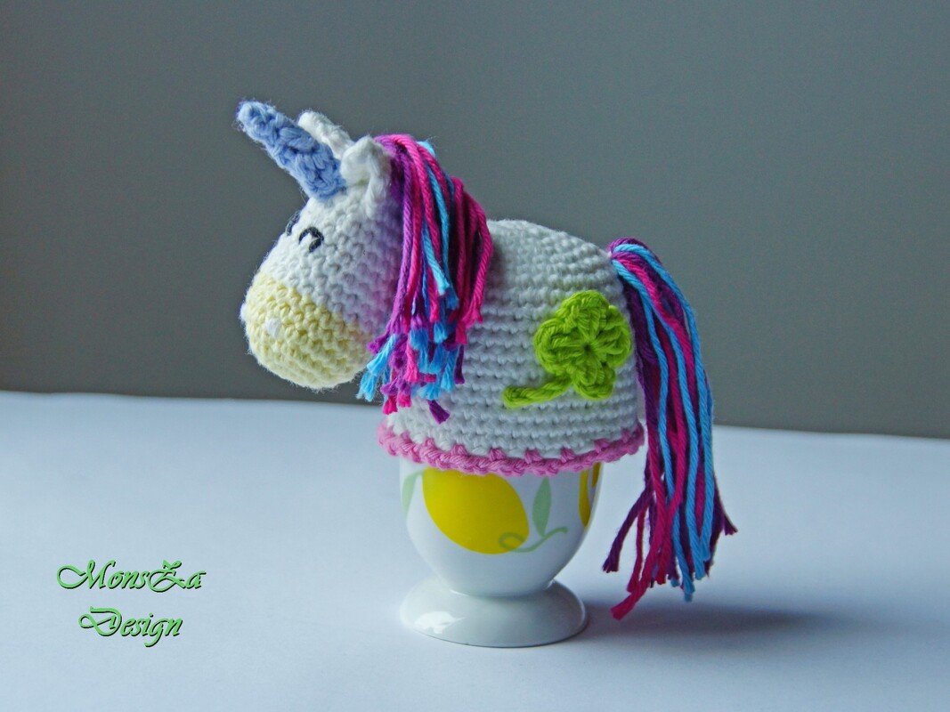 Gehäkelter weißer Einhorn-Eierwärmer mit rosa-blauer Mähne und grünem Kleeblatt auf Eierbecher