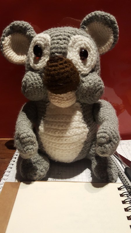 Gehäkelter grauer und weißer Amigurumi-Koala mit brauner Nase und schwarzen Augen auf einem Notizbuch.
