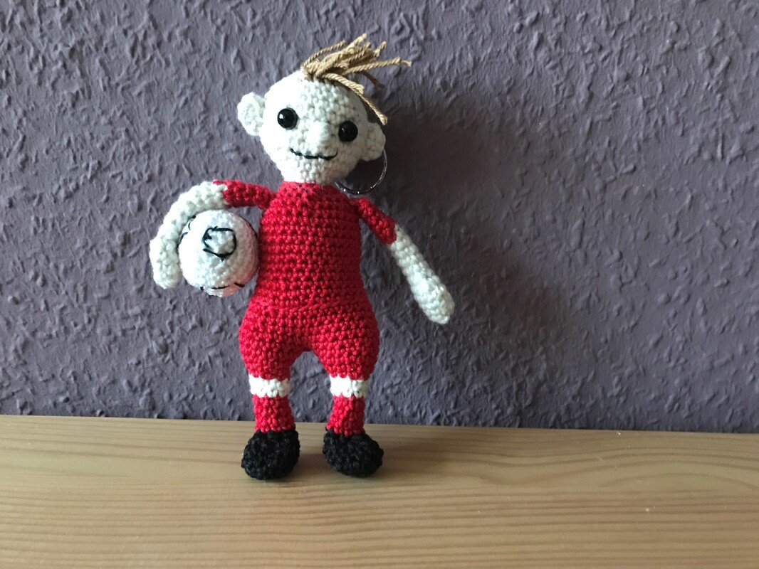 Gehäkelte Amigurumi-Puppe in rotem Outfit, hält einen kleinen Ball, sichtbarer Schlüsselring
