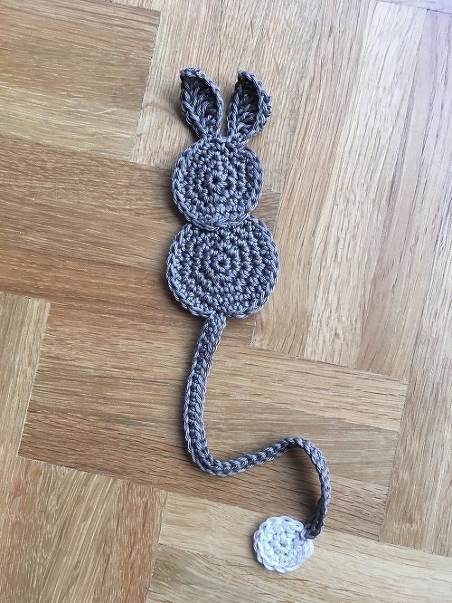 Gehäkeltes graues Hasen-Lesezeichen mit Ohren, langer Kordel und kleinem weißen Häkelkreis auf Holzfußboden