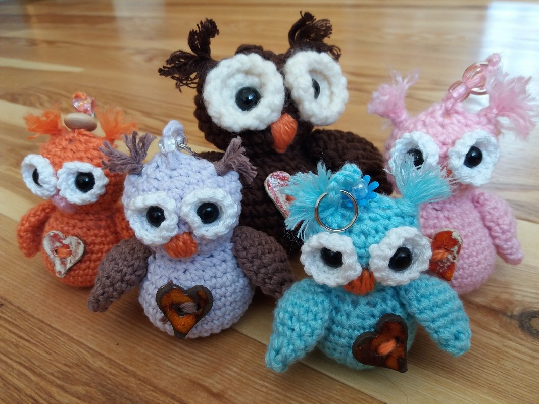 Mehrere kleine gehäkelte Eulen-Amigurumi in verschiedenen Farben mit großen weißen Augen und Herz-Applikationen auf Holzuntergrund