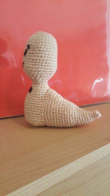 Beige gehäkelte Amigurumi-Figur mit schwarzen gestickten Augen auf Holz vor roter Fläche