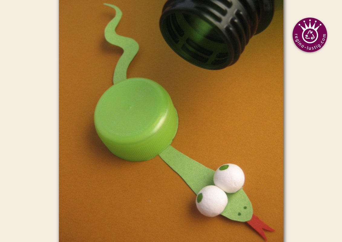 Grüne Schlange aus Flaschendeckel und Papier mit aufgeklebten Augen und roter Zunge auf braunem Hintergrund