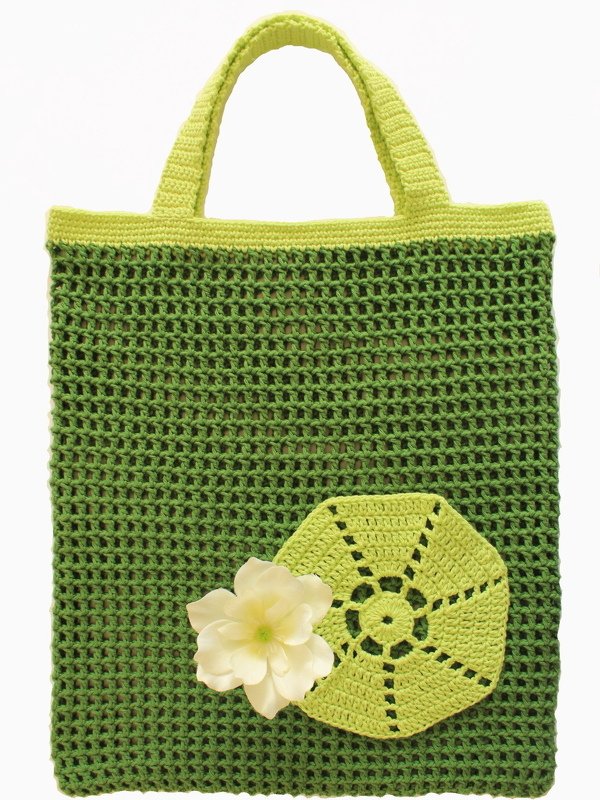Gehäkelte grüne Henkeltasche mit Netzmuster, hellgrünen Henkeln und weißer Blumenapplikation.