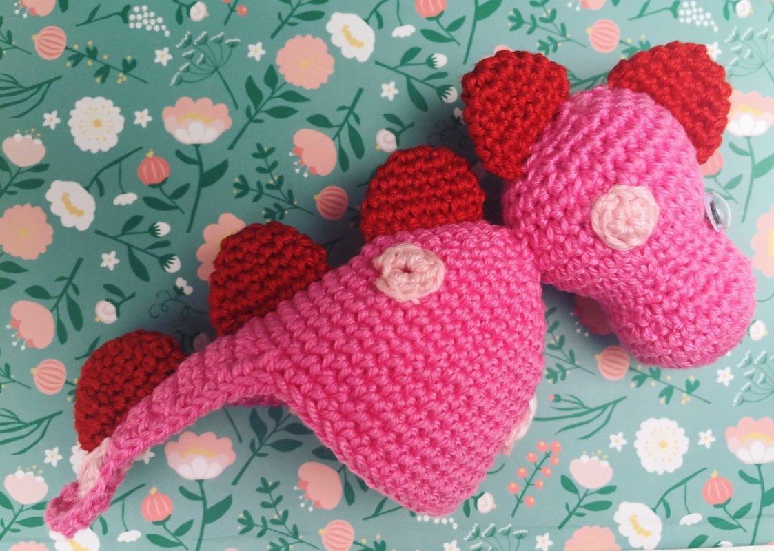 Gehäkelter rosa Drachen-Eierwärmer mit roten Rückenstacheln auf floralem Hintergrund