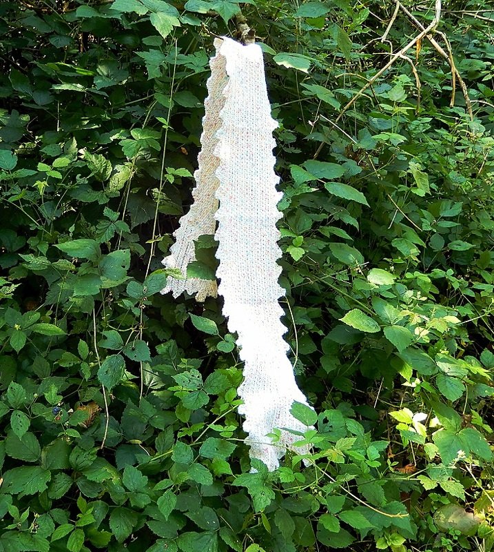 Long white knitted scarf with scalloped edge hanging on green foliage