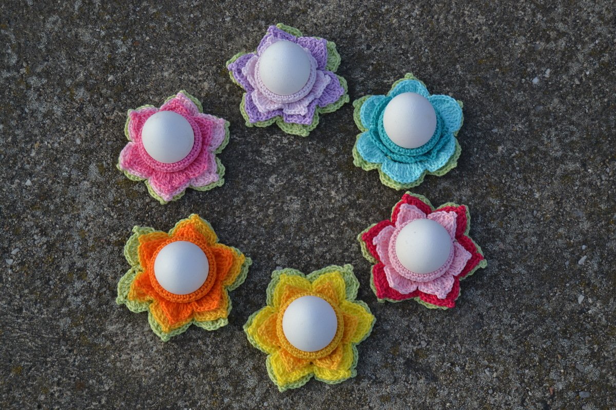 Sechs bunte, gehäkelte Blumen‑Eierwärmer mit weißen Eiern auf grauem Untergrund.