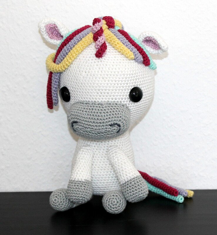 Gehäkeltes weißes Einhorn-Amigurumi mit grauer Schnauze und buntem Schweif, sitzend auf dunkler Fläche vor hellem Hintergrund.