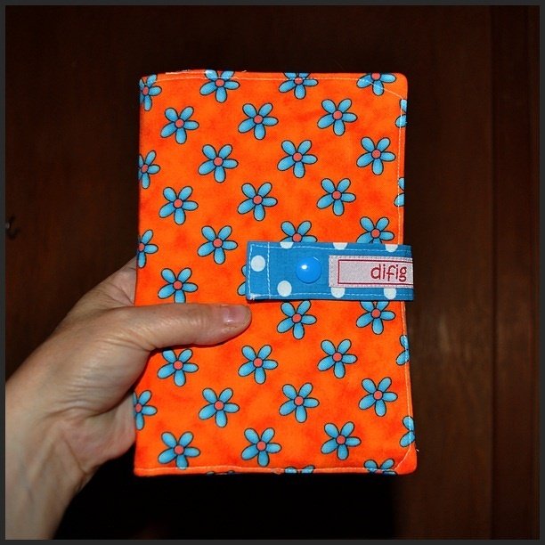 Hand hält orangefarbene Stoffhülle mit blauem Blumenmuster und Lasche mit Druckknopf