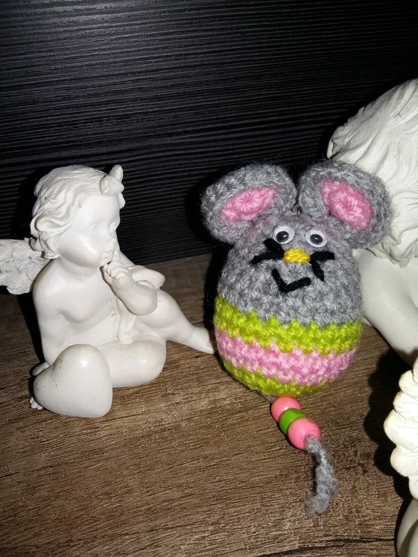 Gehäkelte graue Maus mit rosa Innenohren, rosa‑grünen Streifen, Wackelaugen und Perlen am Schwanz neben weißer Dekofigur