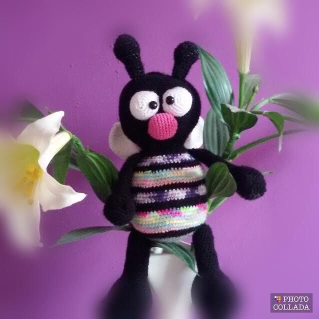 Gehäkelte schwarze Amigurumi-Biene mit buntem gestreiftem Bauch und weißen Flügeln vor lila Hintergrund