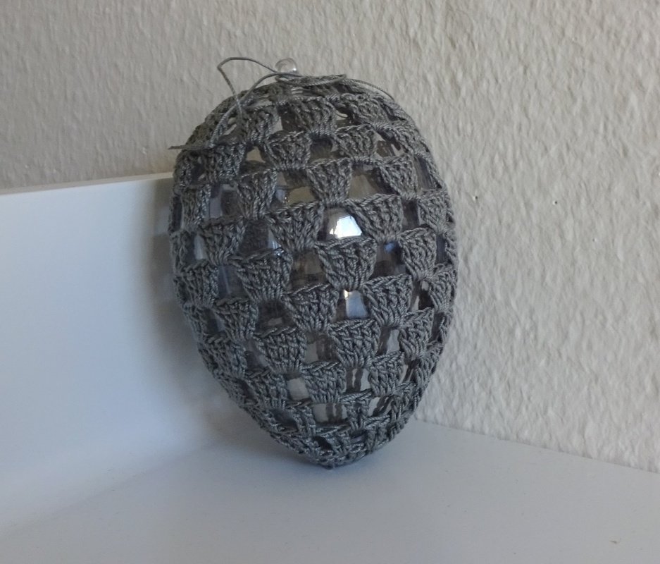 Gehäkeltes graues Ei mit offenem Maschenmuster um ein durchsichtiges Innenobjekt auf weißer Fläche vor heller Wand.
