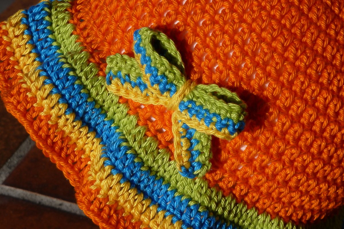 Nahaufnahme einer gehäkelten Kindermütze in Orange mit gelben, blauen und grünen Streifen sowie einer kleinen Schleife.