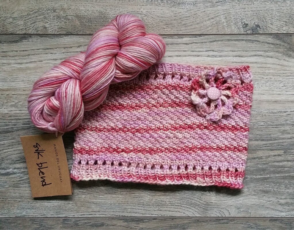 rosa melierter gestrickter Baby-Loop mit Häkelblüte und Garnstrang auf Holzuntergrund