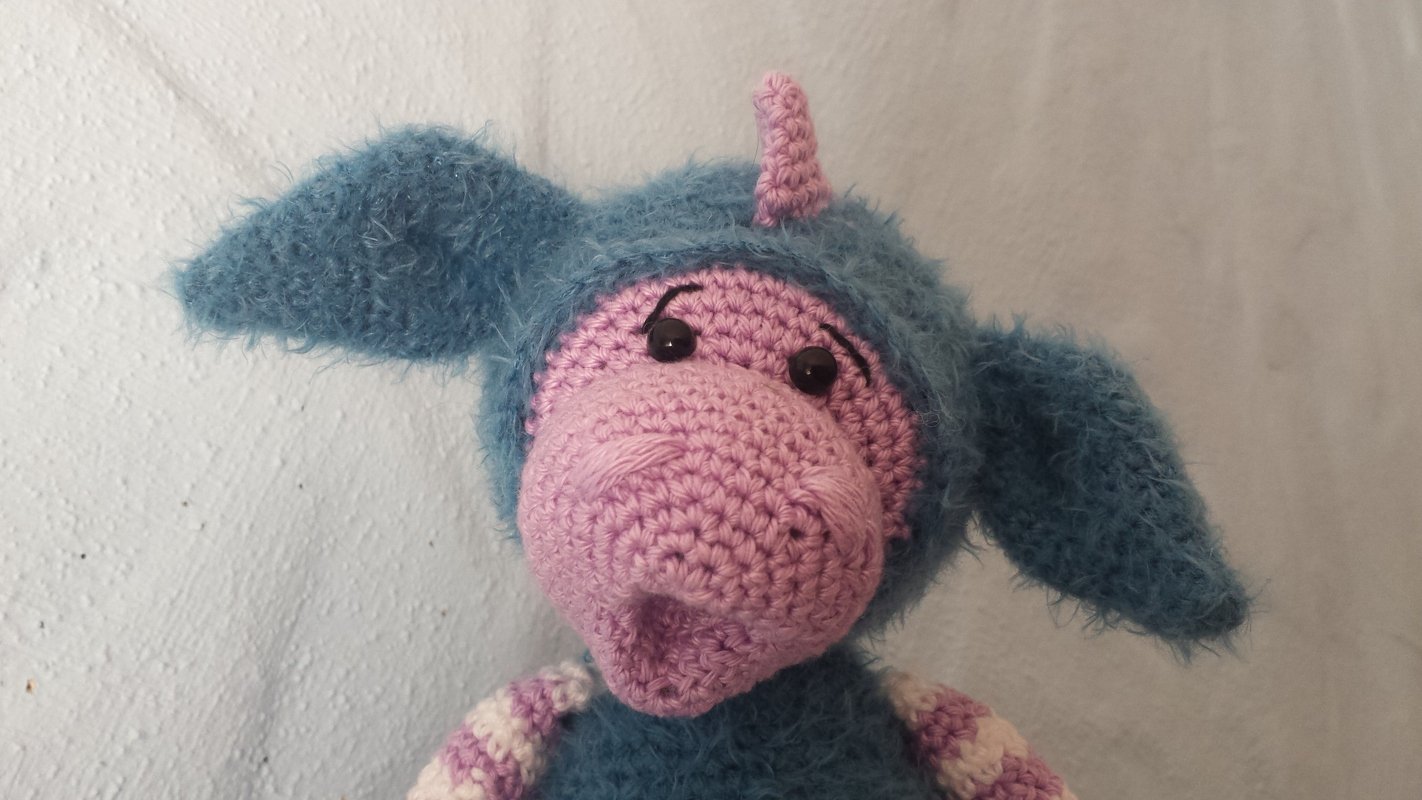 Nahaufnahme eines gehäkelten Amigurumi-Drachenbabys mit flauschiger blauer Kapuze, rosa Schnauze, kleinem Horn und schwarzen Augen.
