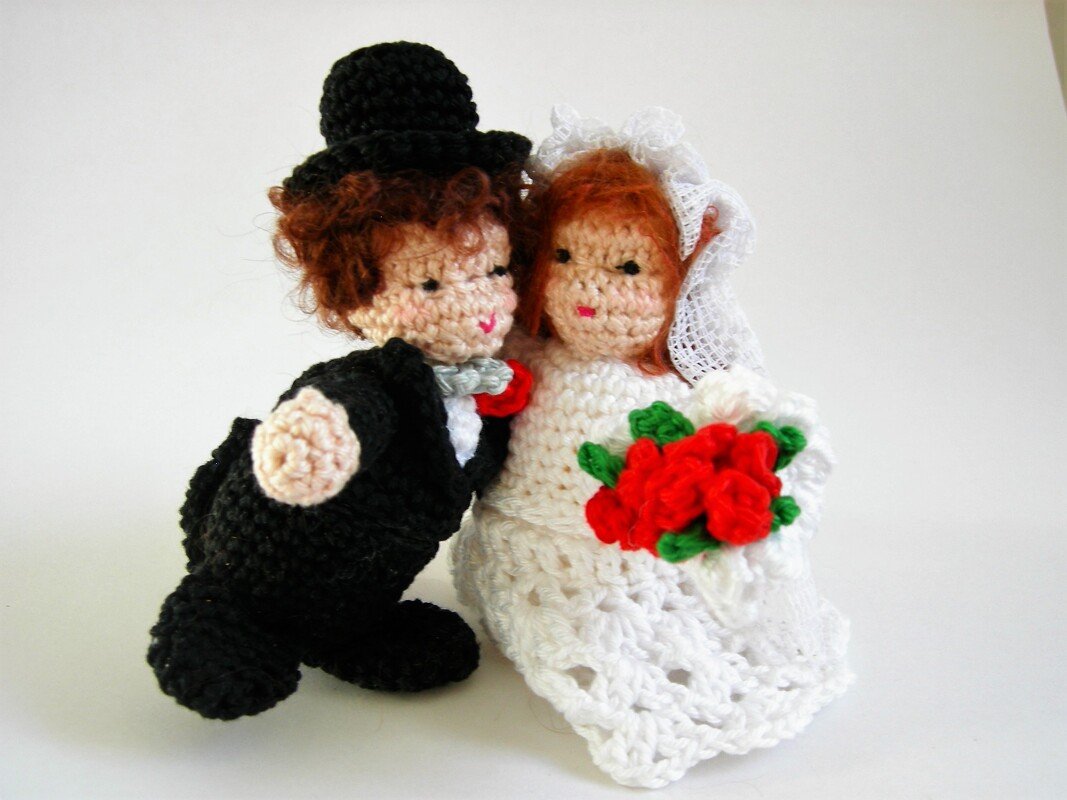 Nahaufnahme eines gehäkelten Brautpaares: Bräutigam in Schwarz mit Zylinder, Braut in Weiß mit Schleier und roten Blumen.