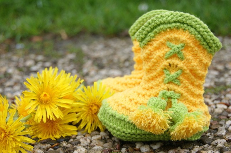 Nahaufnahme gestrickter gelb-grüner Babyschuhe mit Bommeln auf Kiesboden neben Löwenzahn