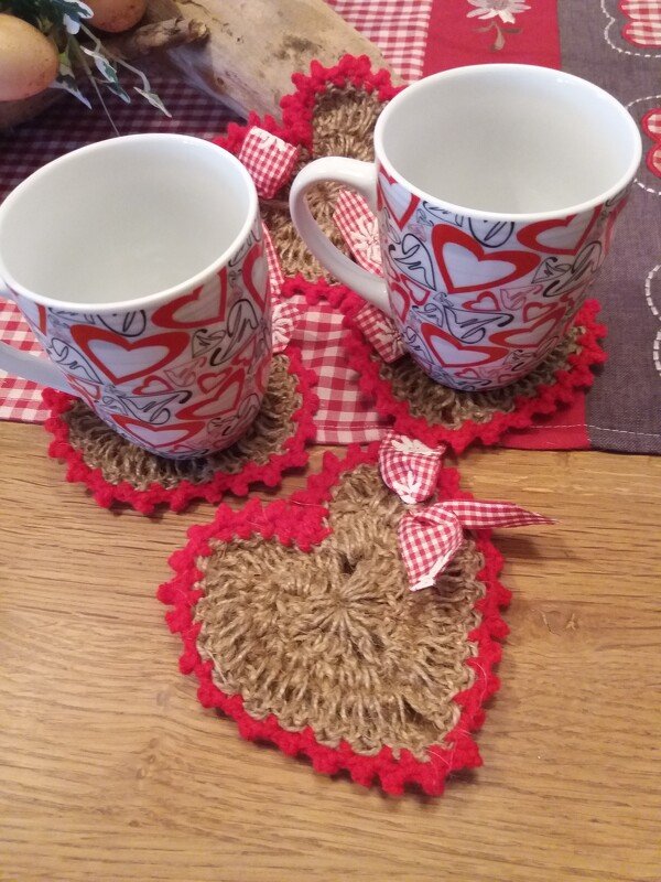 Drei gehäkelte herzförmige Untersetzer in Braun mit rotem Rand und karierten Schleifen, zwei Tassen darauf