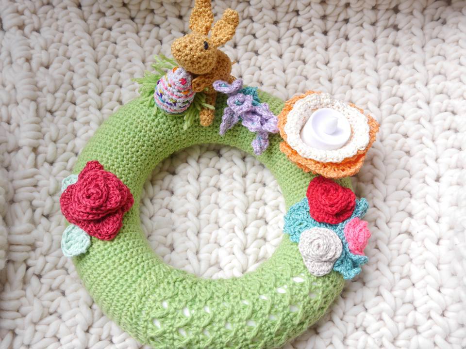 Grüner gehäkelter Kranz auf weißer, grob gestrickter Decke, dekoriert mit braunem Häkelhasen, gestreiftem Osterei und bunten Häkelblüten.