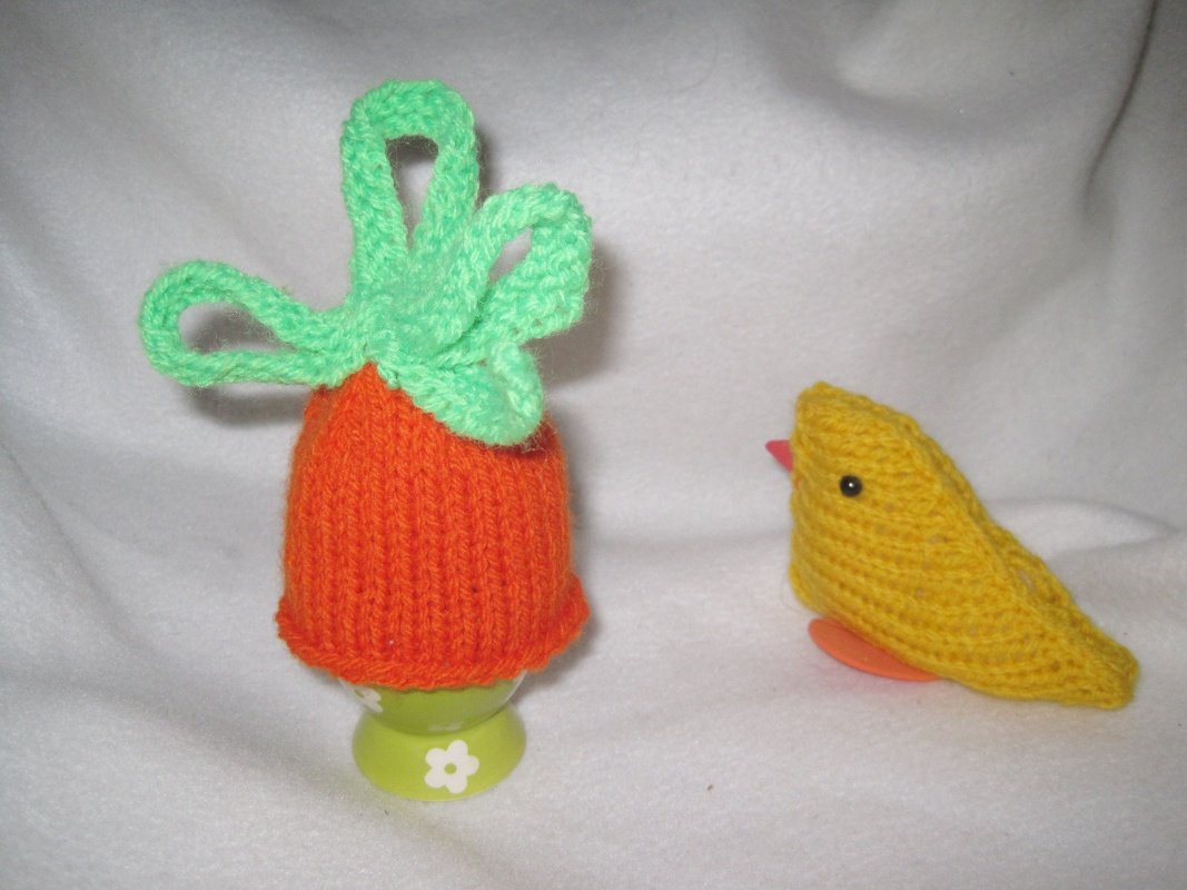 Orange gestrickter Karotten-Eierwärmer mit grünen Blättern auf Eierbecher, daneben gelbes gestricktes Küken