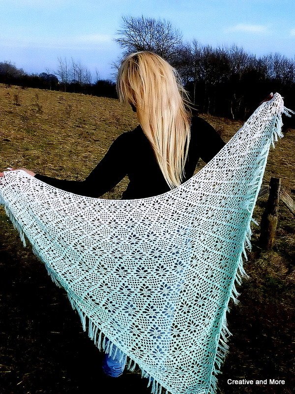 Person seen from behind holding a pale mint crochet triangle shawl with lace openwork and fringe in a field.