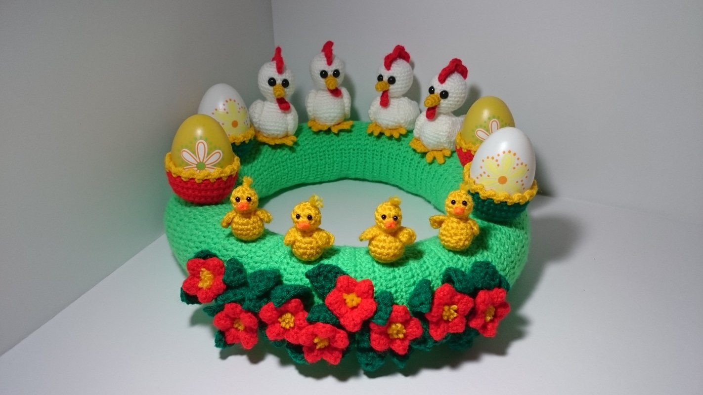 Gehäkelter grüner Osterkranz mit vier weißen Hühnern, vier gelben Küken, bunten Eiern in Körbchen und roten Blüten.